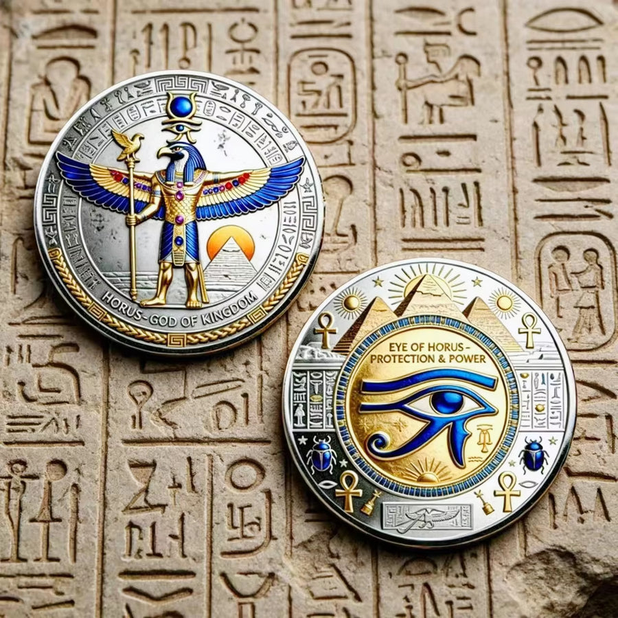 Two circular Egyptian-themed commemorative coins displayed on a beige stone surface with carved hieroglyphs. One coin shows a Horus with blue, gold, and silver details, and the other shows an eye of Horus motif with similar metallic colors.