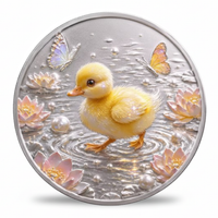 A round silver-toned medallion or coin featuring a yellow duckling standing in water with pink lotus flowers, butterflies, and pearl-like beads, shown on a white background.