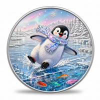 A round silver-toned coin featuring a whimsical winter scene with a penguin wearing a sparkly scarf while sliding on icy water, surrounded by snowy mountains, colorful fish, and a pastel sky.