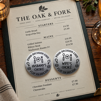 A printed restaurant menu titled 'THE OAK & FORK' laid on a wooden table, with two round metal food decision coins placed on the menu. The coins read 'FOOD DECISION COIN' and 'HUSBAND'S CHOICE' / 'WIFE'S CHOICE' with utensil icons.