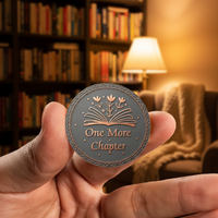 Hand holding a round metal coin with 'One More Chapter' text in a cozy room with books and a lamp.