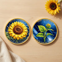 Overhead view of both sunflower coin faces side by side on pale wood, with cream linen at the lower left and a real sunflower at the upper right. The left face shows an open yellow sunflower against a starry blue background with “FOLLOW THE LIGHT” and “BLOOM WITH COURAGE.” The right shows a sunflower bud and green leaves beneath “HOPE · LIGHT · LIFE,” with “GROW TOWARD THE SUN” below, collectible token