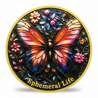 A circular decorative coin featuring a colorful butterfly with orange, pink, and blue wings on a dark background, surrounded by flowers and the text 'Ephemeral Life' in gold at the bottom. The plaque is shown on a clean white background with a soft shadow underneath.