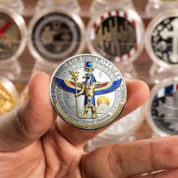 A hand holds a round commemorative coin featuring a gold and blue Egyptian horus with wings, set against a blurred background of other collectible coins in protective cases.