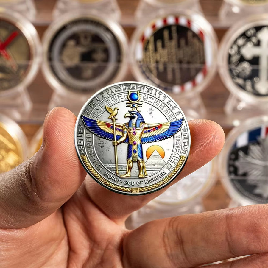 A hand holds a round commemorative coin featuring a gold and blue Egyptian horus with wings, set against a blurred background of other collectible coins in protective cases.
