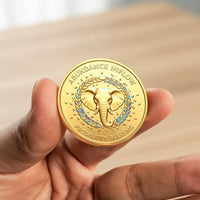 A hand holding a round gold-colored coin featuring an elephant emblem, laurel wreath, and the text 'ABUNDANCE INFLOW' and 'PROSPERITY BLESSING'.