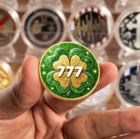 A hand holds a round green and gold 777 slot machine themed coin with a glittery shamrock design, with blurred collectible coins in cases in the background.