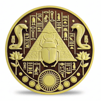 A circular gold-toned coin featuring an Egyptian design with a central pyramid and scarab beetle, flanked by cobras, lotus flowers, and hieroglyphic symbols on a dark brown background.