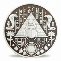 A circular Egyptian-themed coin featuring a raised scarab beetle in front of a pyramid, flanked by two coiled snakes, lotus flowers, and hieroglyphic symbols on a dark brown background with a silver-toned rim.