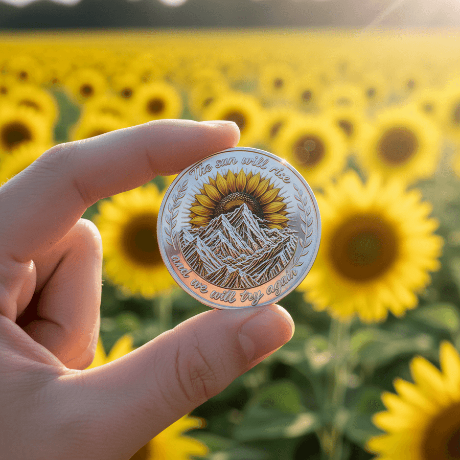 Hand holding mountain sunrise sunflower challenge coin in front of sunflower field collectible token gift

