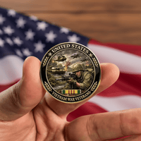 A hand holds a circular United States Vietnam War Veterans commemorative coin in front of a blurred American flag and wooden surface.