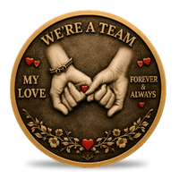 A round decorative coin with a bronze-toned background and raised gold border. The center shows two clasped hands with small red hearts, surrounded by the words “WE'RE A TEAM,” “MY LOVE,” and “FOREVER & ALWAYS,” with floral details along the bottom.